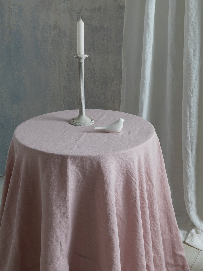 Puede incluir: Un mantel de lino rosa drapeado sobre una mesa redonda con un p&aacute;jaro de cer&aacute;mica blanca y una vela blanca en un candelero de metal gris.