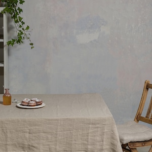 May include: A light brown linen tablecloth covers a table with a clear glass bottle, a plate of small cakes, and a wooden chair with a linen cushion. The table is set against a light gray wall with a vine hanging from the top.