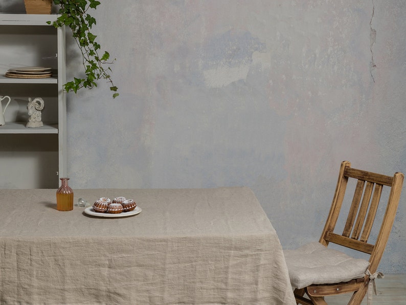 May include: A light brown linen tablecloth covers a table with a clear glass bottle, a plate of small cakes, and a wooden chair with a linen cushion. The table is set against a light gray wall with a vine hanging from the top.