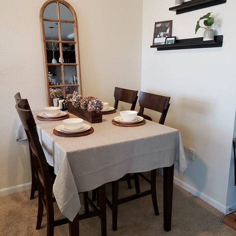 May include: A dining room table set with four chairs, a light grey tablecloth, and white bowls. The table is set with placemats and silverware. There is a vase of flowers in the center of the table.