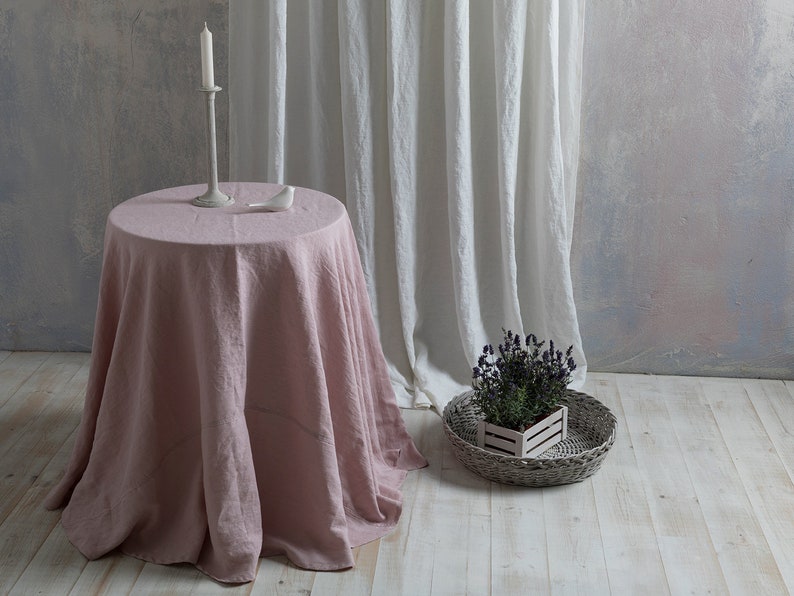 Puede incluir: Un mantel de lino rosa polvoriento drapeado sobre una mesa redonda con una figurita de p&aacute;jaro blanco y un candelero en la parte superior. La mesa est&aacute; delante de una cortina de lino blanco y una pared gris claro.