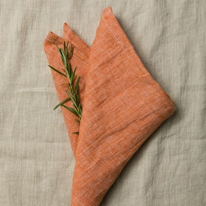 May include: A folded orange linen napkin with a sprig of rosemary on top. The napkin is on a light beige linen background.