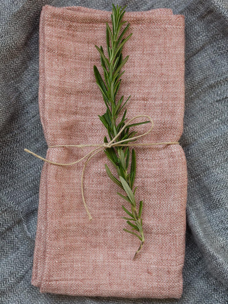 May include: A pink linen napkin folded in half and tied with twine, with a sprig of rosemary on top.