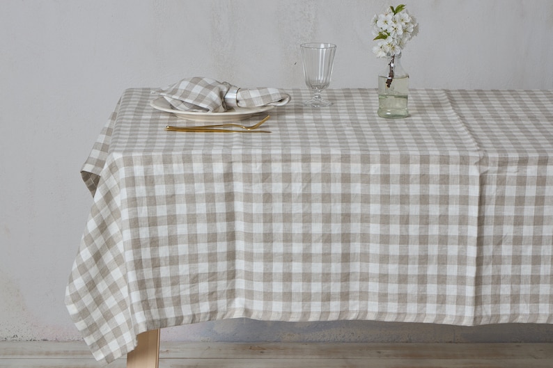 May include: A beige and white checkered linen tablecloth with a white plate, gold silverware, and a glass vase with white flowers on top of a wooden table.