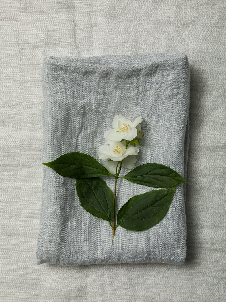 May include: A gray linen throw blanket with a sprig of white jasmine flowers on top.