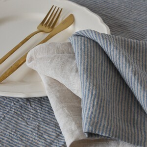 May include: Two linen napkins, one with blue stripes and one plain, are folded on a white plate with gold silverware. The plate is on a blue and white striped tablecloth.