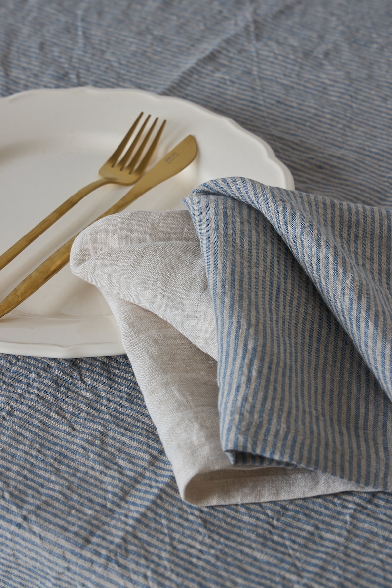 May include: Two linen napkins, one with blue stripes and one plain, are folded on a white plate with gold silverware. The plate is on a blue and white striped tablecloth.