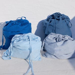 May include: Four drawstring bags in varying shades of blue, ranging from a deep royal blue to a light sky blue. Each bag features a gathered top closure with matching fabric ties, creating a soft, rounded shape for these textile storage pouches.