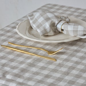 May include: A white and beige gingham tablecloth with a white plate and a linen napkin with a gold napkin ring. A gold fork and knife are placed on the tablecloth.