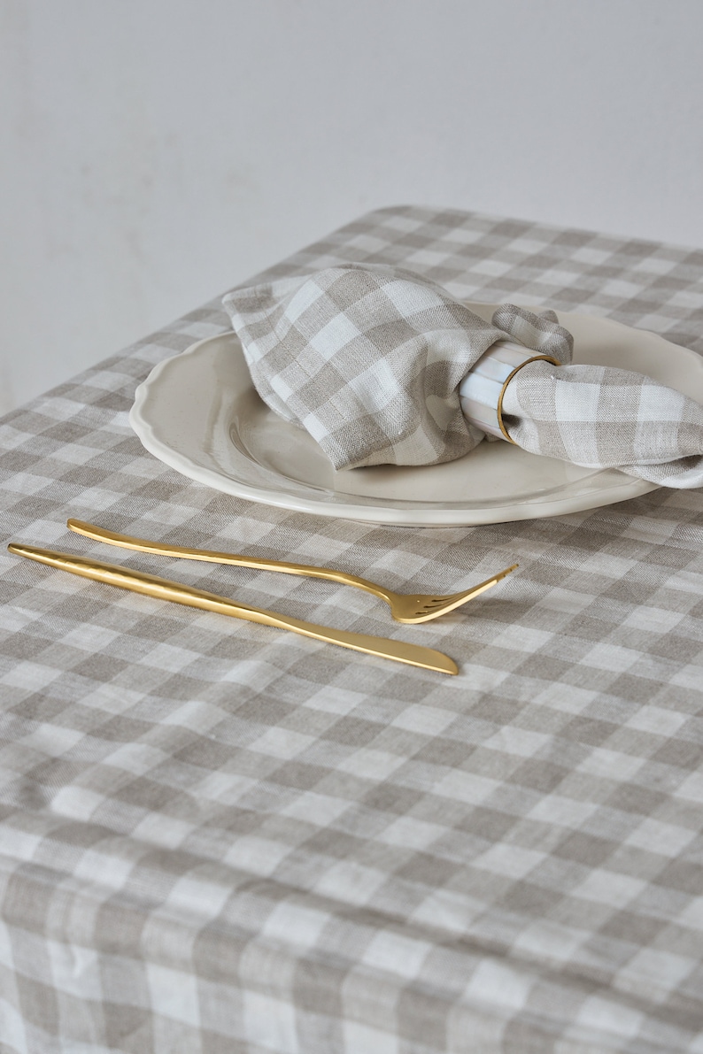 May include: A white and beige gingham tablecloth with a white plate and a linen napkin with a gold napkin ring. A gold fork and knife are placed on the tablecloth.