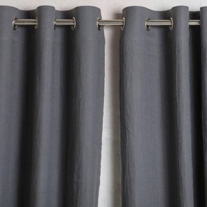 Puede incluir: Un par de cortinas de color gris oscuro con un acabado de tela texturizada. Las cortinas están suspendidas de una barra de metal plateado mediante ojales metálicos circulares espaciados, creando pliegues verticales en los paneles.