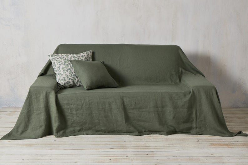 Op de afbeelding: Een groene linnen sofa deken met twee kussens, een met een bloemenpatroon en een effen groen.