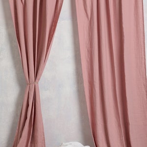 May include: A pair of dusty rose linen curtains hanging from a wooden rod. The curtains are gathered at the top and have a relaxed, casual look. A woven basket with a white cloth inside sits in front of the curtains.