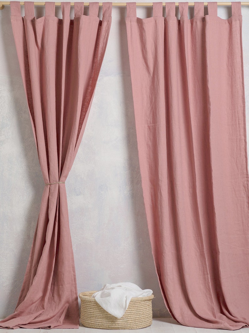 May include: A pair of dusty rose linen curtains hanging from a wooden rod. The curtains are gathered at the top and have a relaxed, casual look. A woven basket with a white cloth inside sits in front of the curtains.