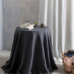 May include: A dark gray linen tablecloth draped over a round table with a stack of books, two small potted plants, and a ball of yarn on top. The table is in front of a white linen curtain.