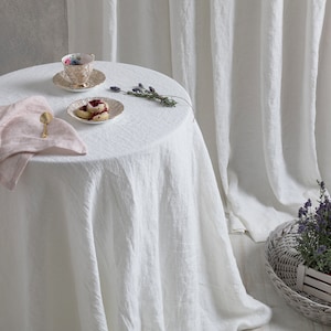 May include: A white linen tablecloth with a pink linen napkin, a teacup and saucer, a plate with scones, and a sprig of lavender on a white round table.