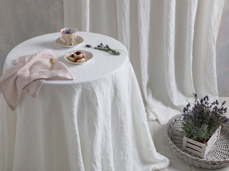 May include: A white linen tablecloth with a pink linen napkin, a teacup and saucer, a plate with scones, and a sprig of lavender on a white round table.