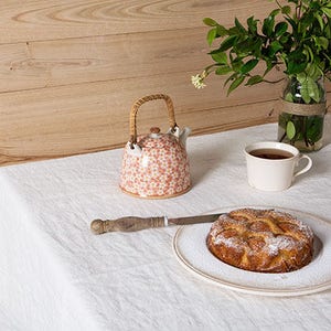 May include: A white tablecloth with a teapot, a cup of tea, and a cake on a plate. The teapot is white with a floral pattern and a wicker handle. The cup is white with a handle. The cake is round and has a brown crust with a white glaze. The cake is dusted with powdered sugar.
