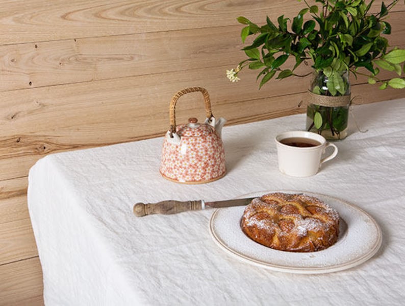 May include: A white tablecloth with a teapot, a cup of tea, and a cake on a plate. The teapot is white with a floral pattern and a wicker handle. The cup is white with a handle. The cake is round and has a brown crust with a white glaze. The cake is dusted with powdered sugar.