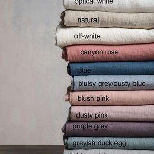 May include: A stack of linen fabric swatches in various shades of grey, pink, blue, and white. The swatches are labeled with their color names, including "optical white", "natural", "off-white", "canyon rose", "blue", "bluisy grey/dusty blue", "blush pink", "dusty pink", "purple grey", "greyish duck egg", "light grey/silver grey", and "dark grey".