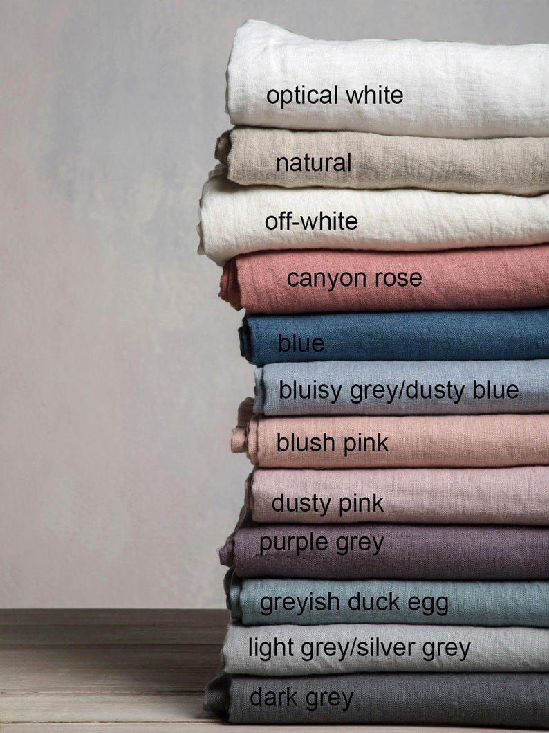 May include: A stack of linen fabric swatches in various shades of grey, pink, blue, and white. The swatches are labeled with their color names, including "optical white", "natural", "off-white", "canyon rose", "blue", "bluisy grey/dusty blue", "blush pink", "dusty pink", "purple grey", "greyish duck egg", "light grey/silver grey", and "dark grey".