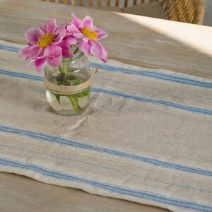 May include: A clear glass jar with a brown twine bow around the top, filled with pink peonies and water. The jar is sitting on a white and blue striped table runner.
