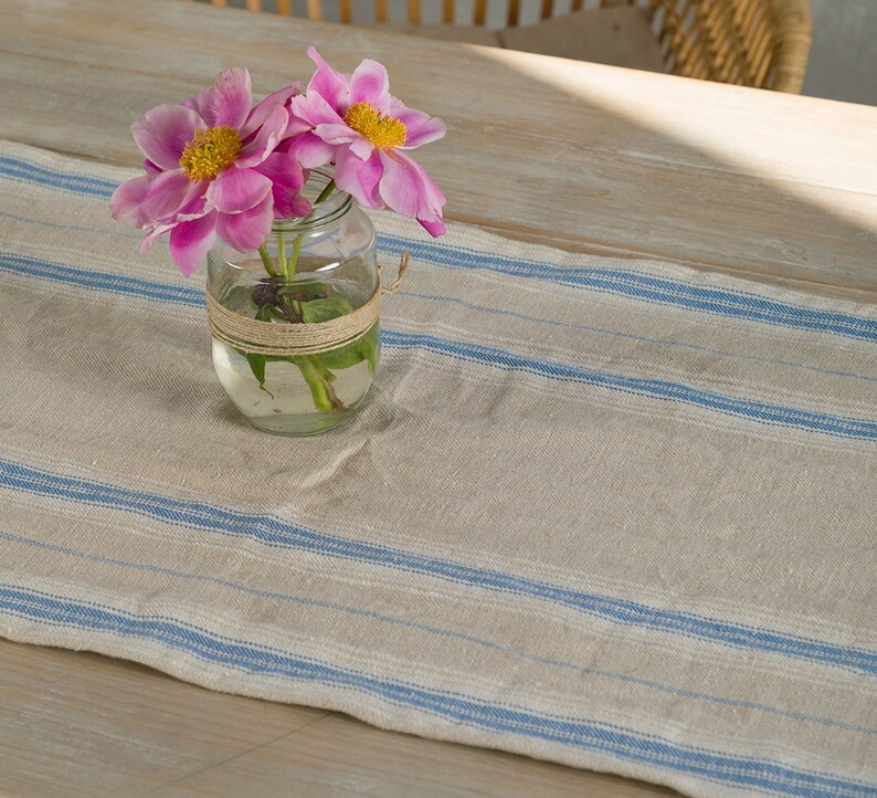May include: A clear glass jar with a brown twine bow around the top, filled with pink peonies and water. The jar is sitting on a white and blue striped table runner.