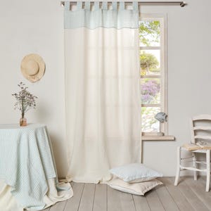 May include: White linen curtains with blue and white striped trim, hanging in a room with a window, a table with a blue and white striped tablecloth, a chair, and a hat on the wall.