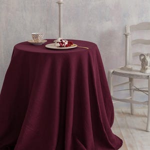 May include: A round table covered with a deep burgundy linen tablecloth. A plate with dessert and berries sits on the table, along with a teacup and a tall candle holder with a lit candle. A white chair is in the background.