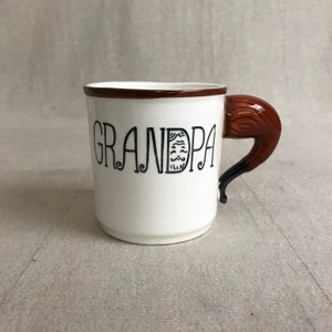 May include: White ceramic mug with a brown handle shaped like a horse's head. The mug says "GRANDPA" in black lettering.