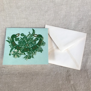 Pode incluir: Um cartão azul claro com um design verde e dourado apresentando um motivo floral estilizado com o texto "In Vita Ut In Morte" em letras douradas.