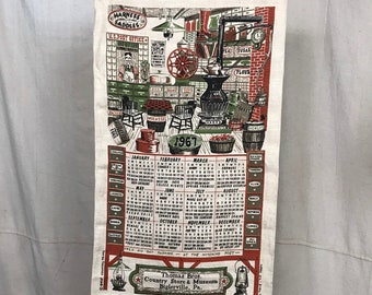 May include: Vintage 1967 calendar featuring a detailed illustration of a country store interior. The artwork includes shelves stocked with goods, a U.S. Post Office, and a calendar grid. The calendar is printed on a cream-colored fabric and is suspended by a wooden rod.