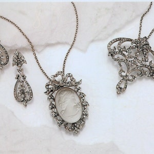 K&ouml;nnte beinhalten: Ein Set aus silberfarbenem Schmuck mit einem Kameenanh&auml;nger mit einem wei&szlig;en Kamee, einem Paar baumelnden Ohrringen in Tropfenform und einer Brosche mit floralem Design.