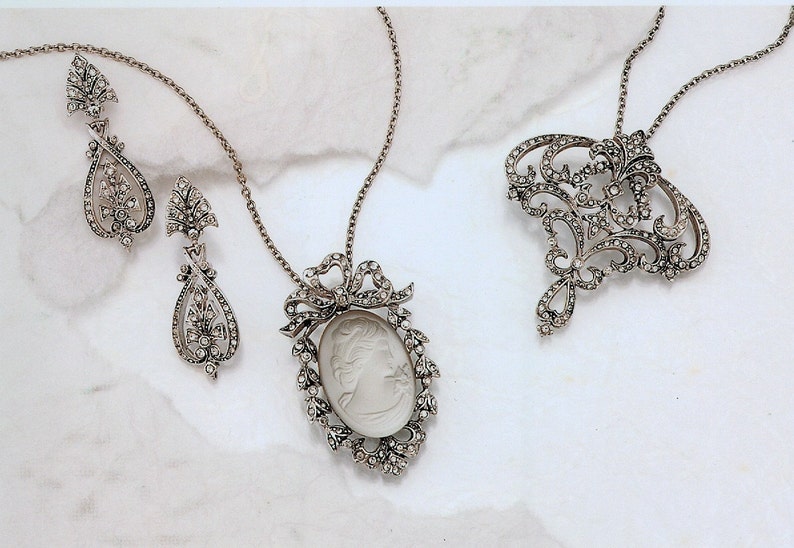 K&ouml;nnte beinhalten: Ein Set aus silberfarbenem Schmuck mit einem Kameenanh&auml;nger mit einem wei&szlig;en Kamee, einem Paar baumelnden Ohrringen in Tropfenform und einer Brosche mit floralem Design.