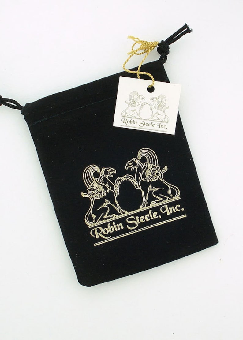 K&ouml;nnte beinhalten: Eine schwarze Samt-Zugschnur-Tasche mit einem goldenen Logo, das "Robin Steele, Inc." lautet und zwei stilisierte Greife zeigt. Die Tasche wird von einem wei&szlig;en Etikett mit dem gleichen Logo begleitet.