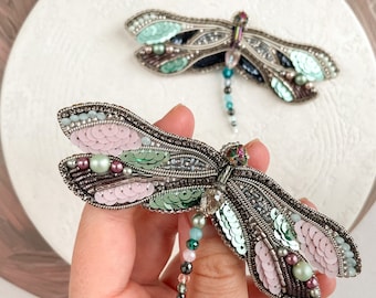 May include: Two beaded dragonfly brooches. The brooches feature silver-toned metalwork, iridescent sequins, and colorful beads in shades of pink, green, and blue. One brooch is held in a hand, while the other rests on a white surface.