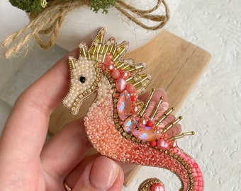 Puede incluir: Broche artesanal de caballito de mar en tonos coral, melocotón y dorado. El diseño con cuentas presenta cristales iridiscentes, perlas y detalles dorados. Un accesorio único para realzar cualquier atuendo.