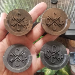 May include: Four round coasters with a raised design. The coasters are made of a dark brown and black material. The design features a cross with the letters "C" on each end and a small lizard in the center.