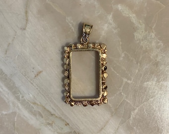May include: A gold-colored rectangular pendant with a textured, beaded border. The pendant has a loop at the top for a chain. The center is open, designed to hold a stone or photo. The pendant is displayed on a marble surface.