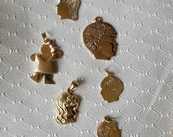 May include: Six gold-tone pendants with profile images of children. The pendants are on a white fabric background.
