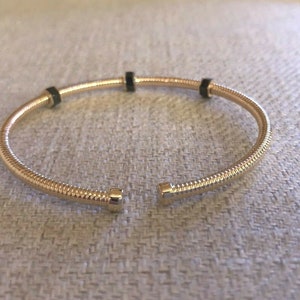 Puede incluir: Un brazalete de oro con cuatro acentos negros. El brazalete está abierto y tiene una bisagra de resorte.