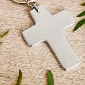 May include: Silver cross keychain with a silver key ring. The keychain has the text "Céline Dupré Bijoux" on it.