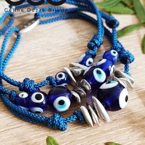 Peut inclure: Un bracelet bleu et argent avec plusieurs charmes œil maléfique. Le bracelet est fait d'un cordon bleu et a des perles et des breloques argentées. Les charmes œil maléfique sont bleus avec un œil blanc au centre. Le bracelet est noué et porte le texte "Céline Dupré Bijoux" sur l'étiquette.
