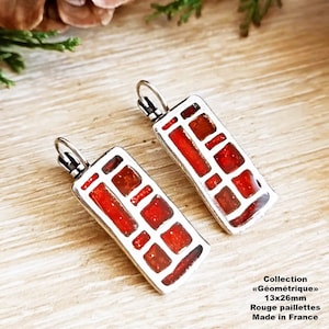 May include: A pair of silver earrings with red geometric designs. The earrings are made with red glitter and measure 1.3 x 2.6 inches. The earrings are part of the "Geometrique" collection and are made in France.