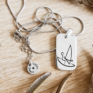 Peut inclure: Collier en argent avec un pendentif rectangulaire représentant un dessin au trait d'une personne faisant de la planche à voile. Le pendentif est attaché à une chaîne en argent avec un petit charme rond qui dit "Celine Dupre" avec une étoile.
