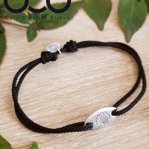 May include: A black cord bracelet with a silver charm featuring a seashell design. The charm is oval-shaped and has a raised seashell design. The bracelet is tied with a knot at the end.