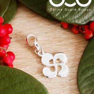 May include: Silver charm with a stylized flower design. The charm has a loop at the top for attaching to a bracelet or necklace. The charm is on a wooden surface with green leaves and red berries.