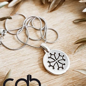 Può includere: Una collana con pendente in argento con un pendente circolare con un design floreale nero stilizzato. La collana poggia su una superficie di legno, con foglie secche sullo sfondo. Il marchio "Céline Dupré Bijoux" è visibile.