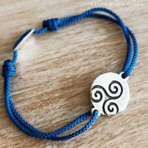 Puede incluir: Una pulsera de cordón azul con un dije circular plateado que presenta un diseño de espiral triskelion en negro. La pulsera incluye nudos deslizantes ajustables y una pequeña etiqueta plateada. El texto "cdb Céline Dupré Bijoux" es visible.