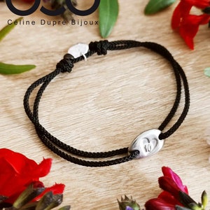 Peut inclure: Un bracelet en cordon noir avec un charme ovale argenté représentant un cœur avec le chiffre 10 à l'intérieur. Le bracelet est sur une surface en bois avec des fleurs rouges et violettes.